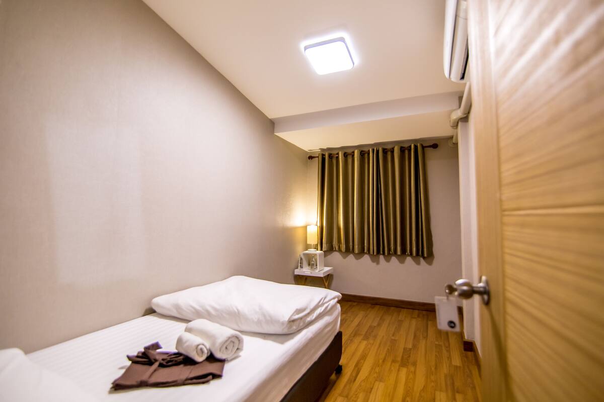 A hotel room with a bed, a bedside table with a lamp, a window with curtains, and a door.
