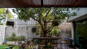 Uma área externa com mesas e cadeiras, cercada por uma árvore e plantas pendentes.