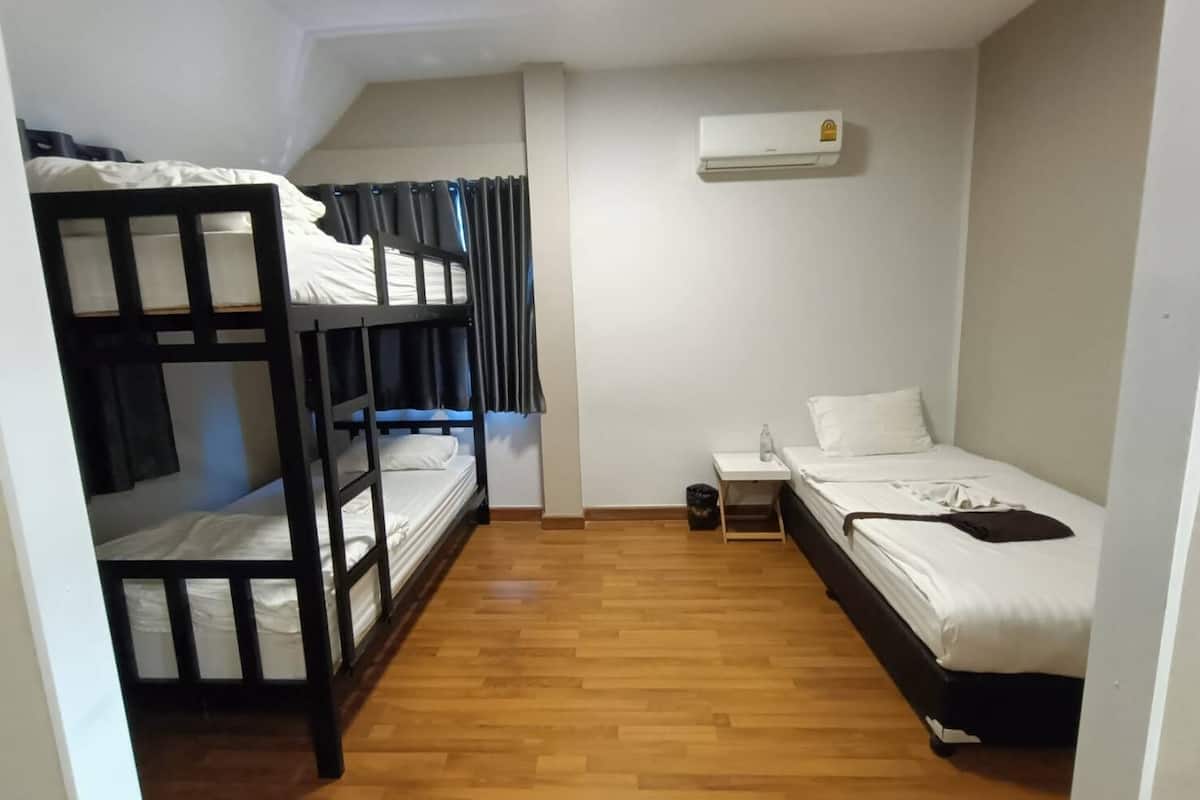 A dormitory room with bunk beds, a single bed, a small table with a bottle, and an air conditioning unit.