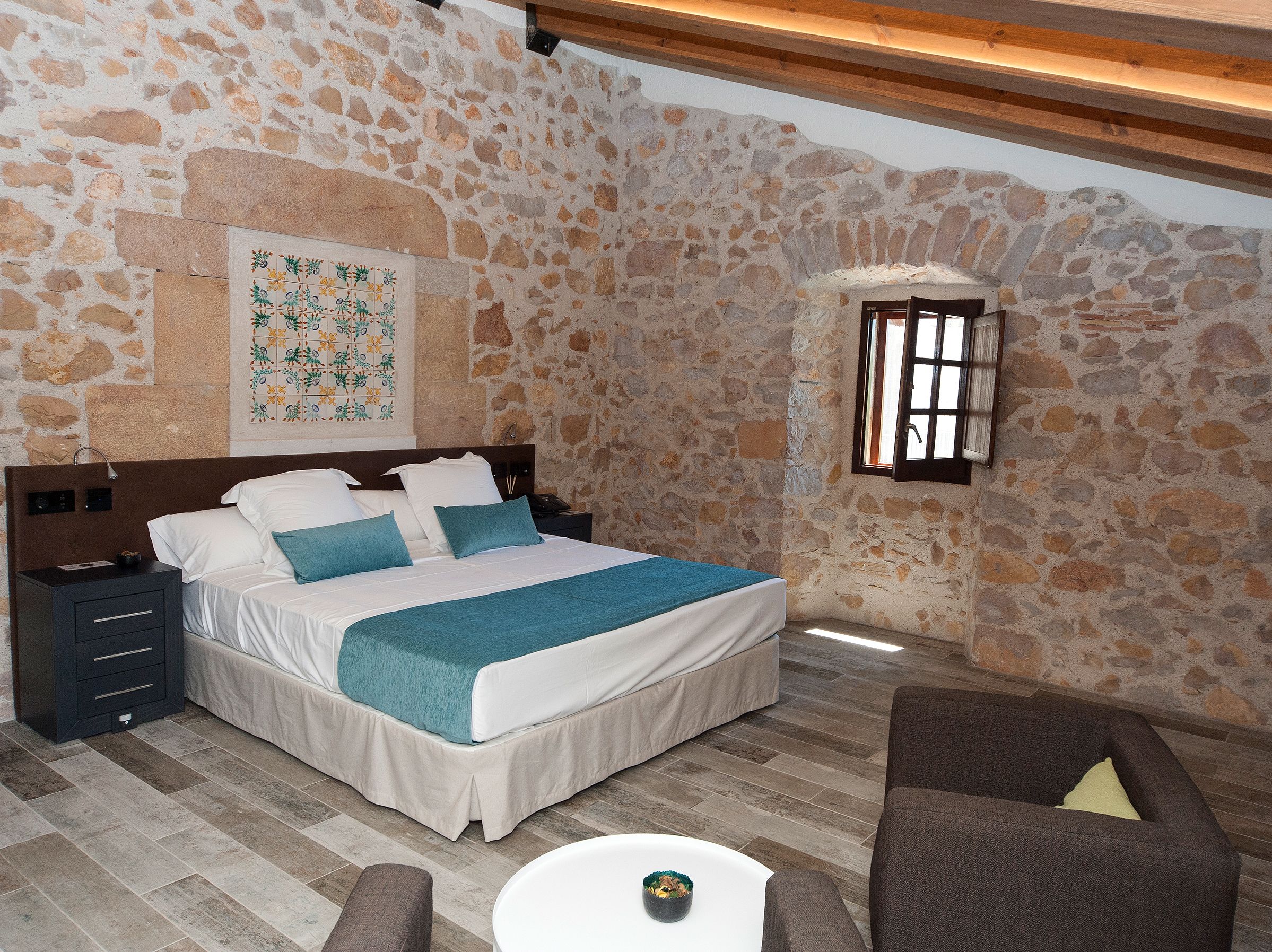 Quarto com parede de pedra, cama com roupa de cama branca e detalhes em azul-esverdeado, mesa de cabeceira, pequena mesa com uma tigela e janela com grades.