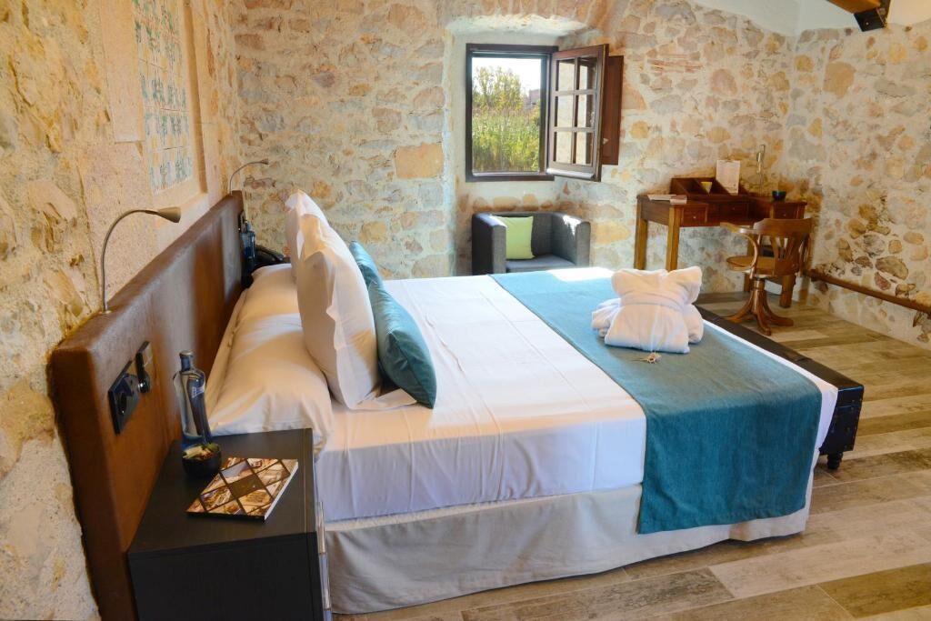 Un dormitorio con pared de piedra, cama con edredón azul y blanco, mesita de noche con lámpara y ventana con vista a vegetación.