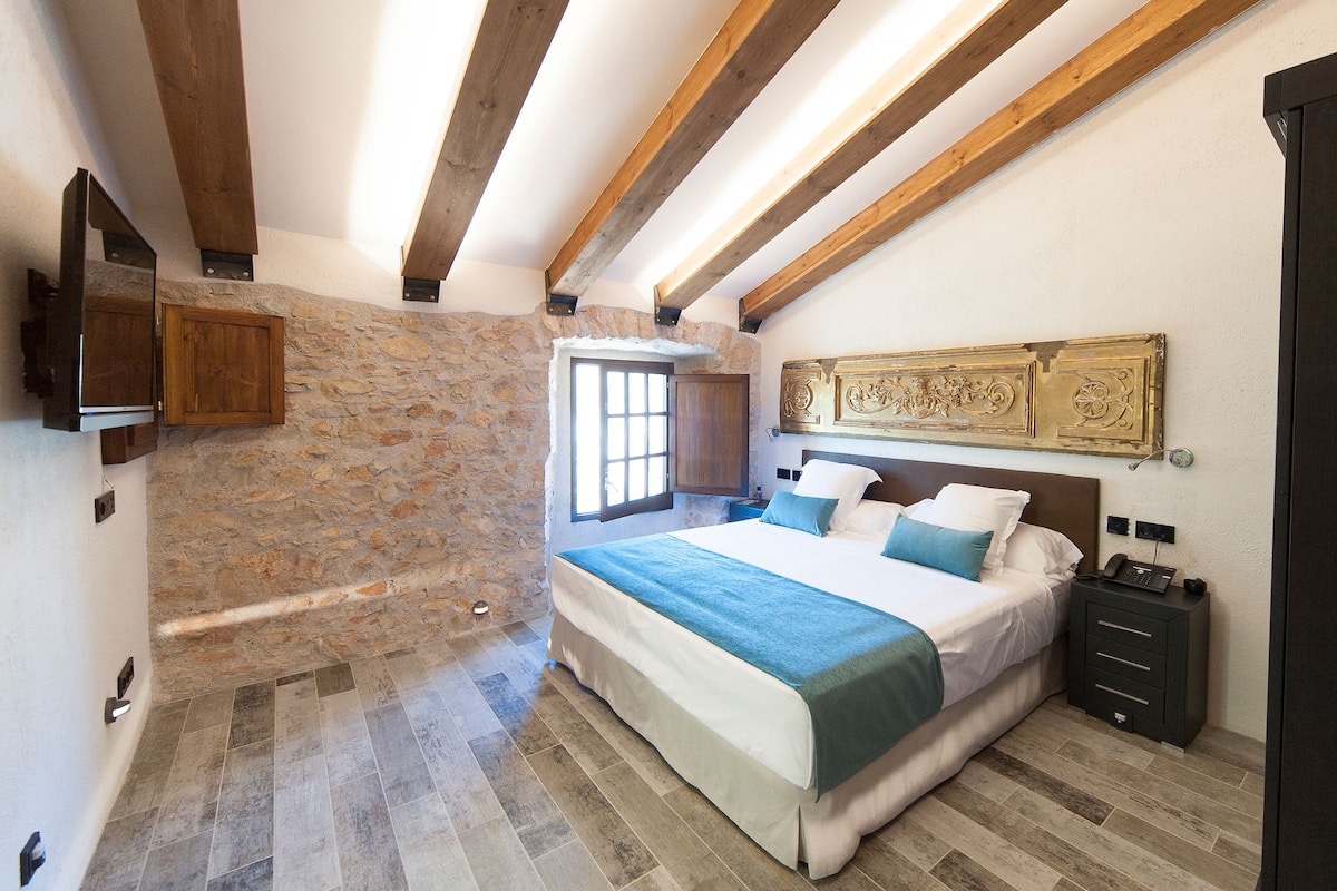 A bedroom with a stone wall, wooden beams, a bed with a blue and white bedspread, a nightstand, and a mounted TV.