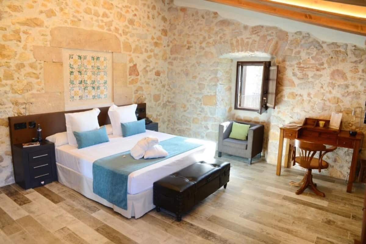 A bedroom with a stone wall, a bed with a blue blanket, a wooden desk, and a chair.
