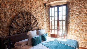 A bedroom with a stone wall, a metal headboard, a large window, and a bed with pillows and a blanket. - Hotel Molí de l'Escala (L'Escala)