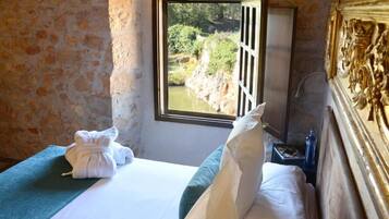 Una habitación con pared de piedra, cama con ropa de cama blanca, toalla verde y ventana con vista a vegetación.