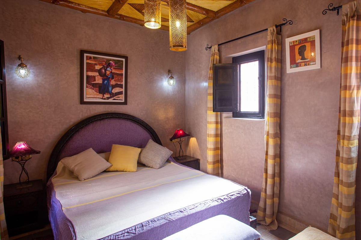 A bedroom with a bed, a nightstand, a lamp, a window with curtains, and wall art.