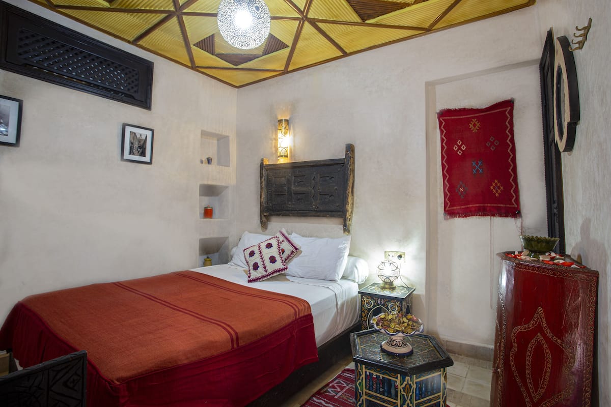 A bedroom with a bed, a nightstand, a red decorative item, and a wall-mounted shelf.