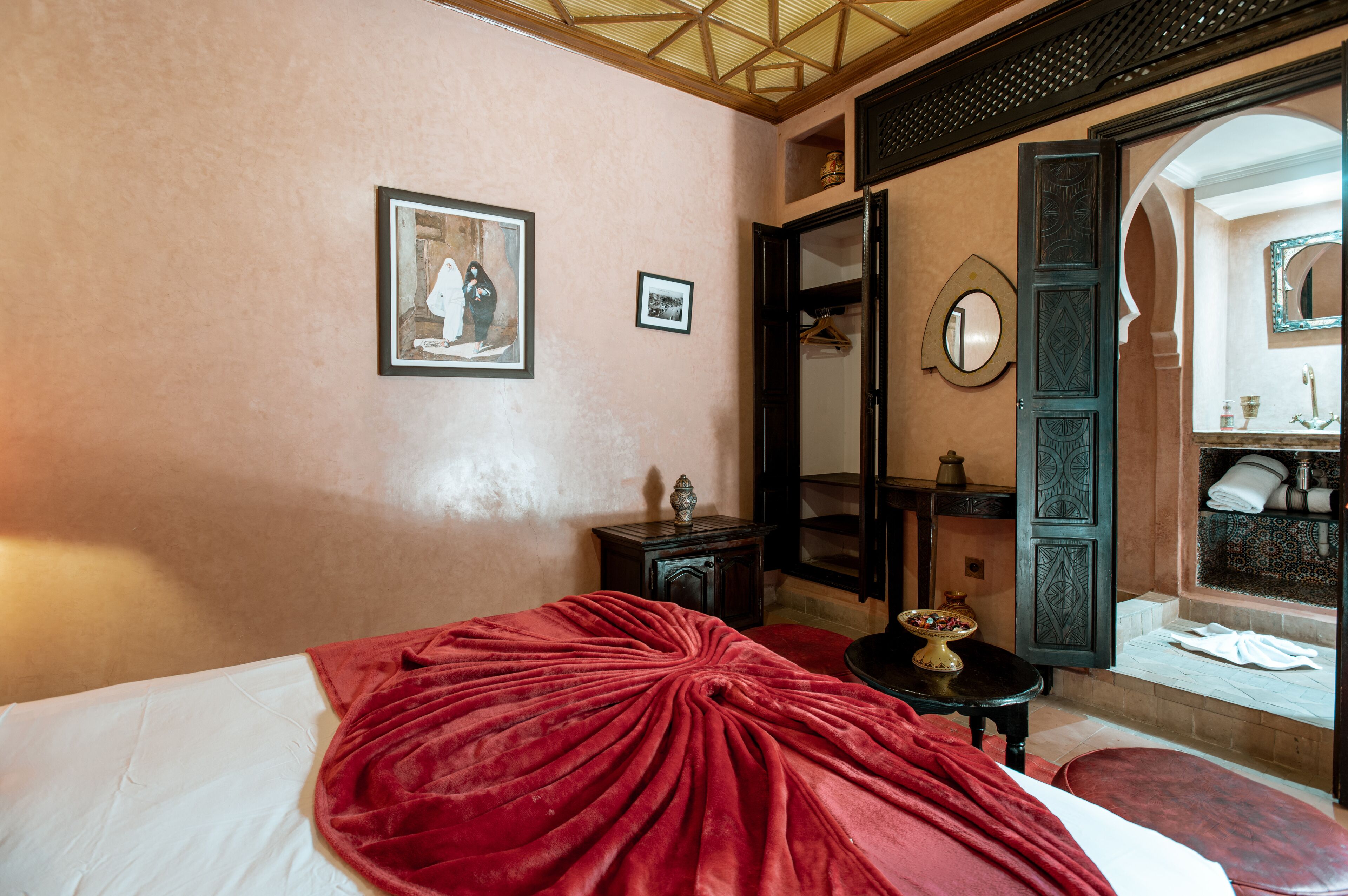 A hotel room with a bed, a red blanket, a small table with a bowl, a mirror, and a bathroom visible through an open door.