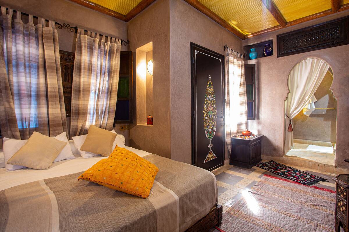 A hotel room with a bed, curtains, a door with a decorative design, a small table with a vase, and a mirror.