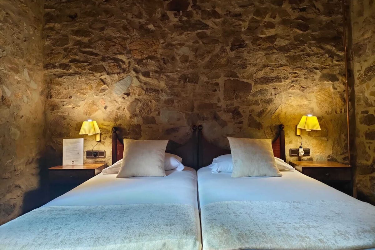 A room with two beds, stone walls, and two bedside tables with lamps.