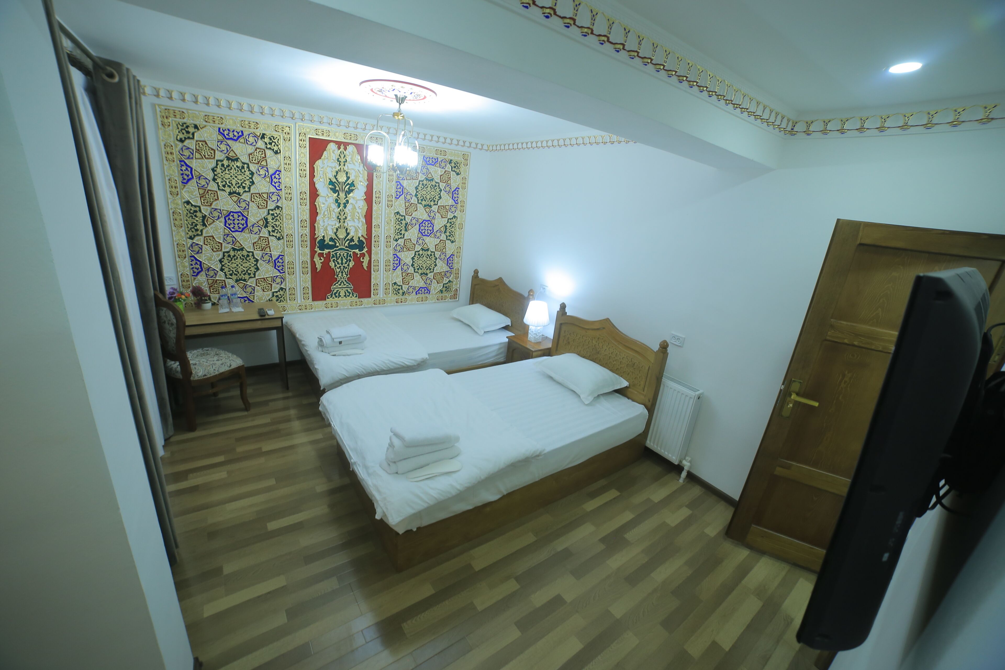 A hotel room with two beds, a wooden floor, a decorative wall tapestry, a wooden door, and a television.