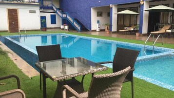 A swimming pool with a glass table and wicker chairs, surrounded by a grassy area and a blue building.