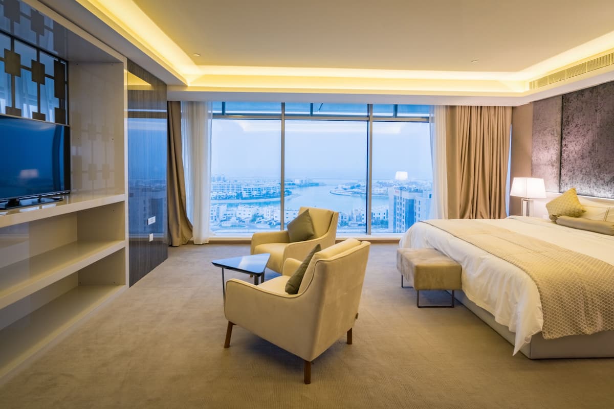 Quarto de hotel com uma cama grande, um sofá, uma TV em uma estante e vista para a cidade através de grandes janelas.