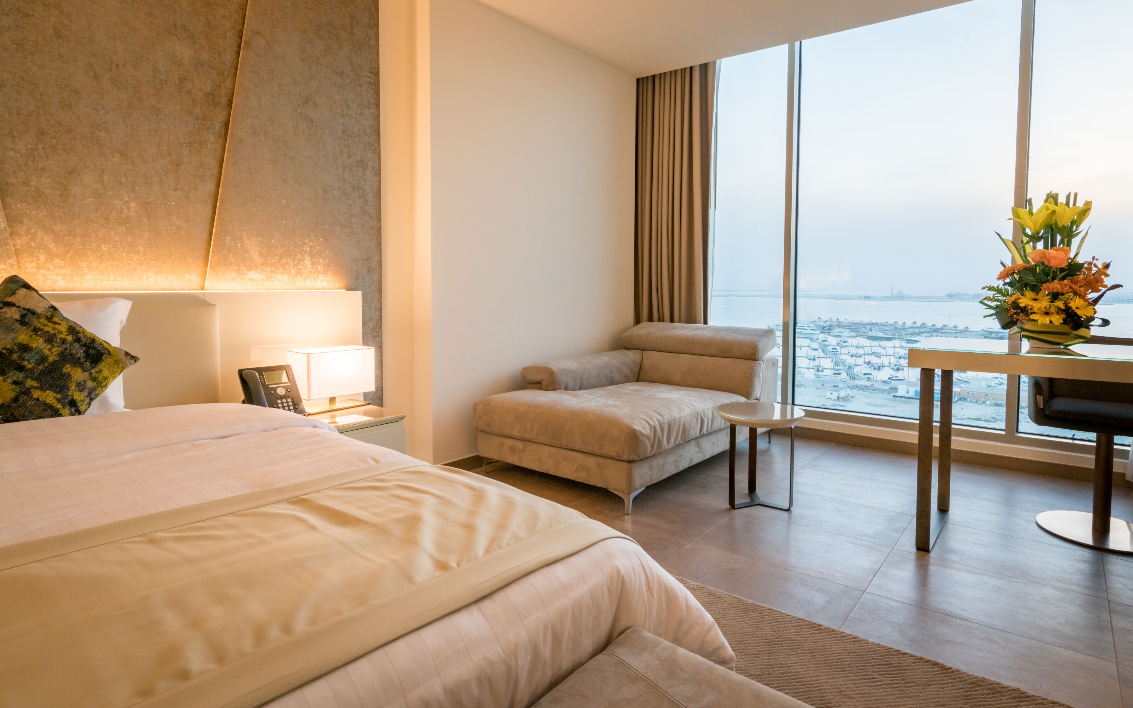 Habitación de hotel con una cama grande, una silla, un escritorio y vistas de la ciudad a través de amplias ventanas.