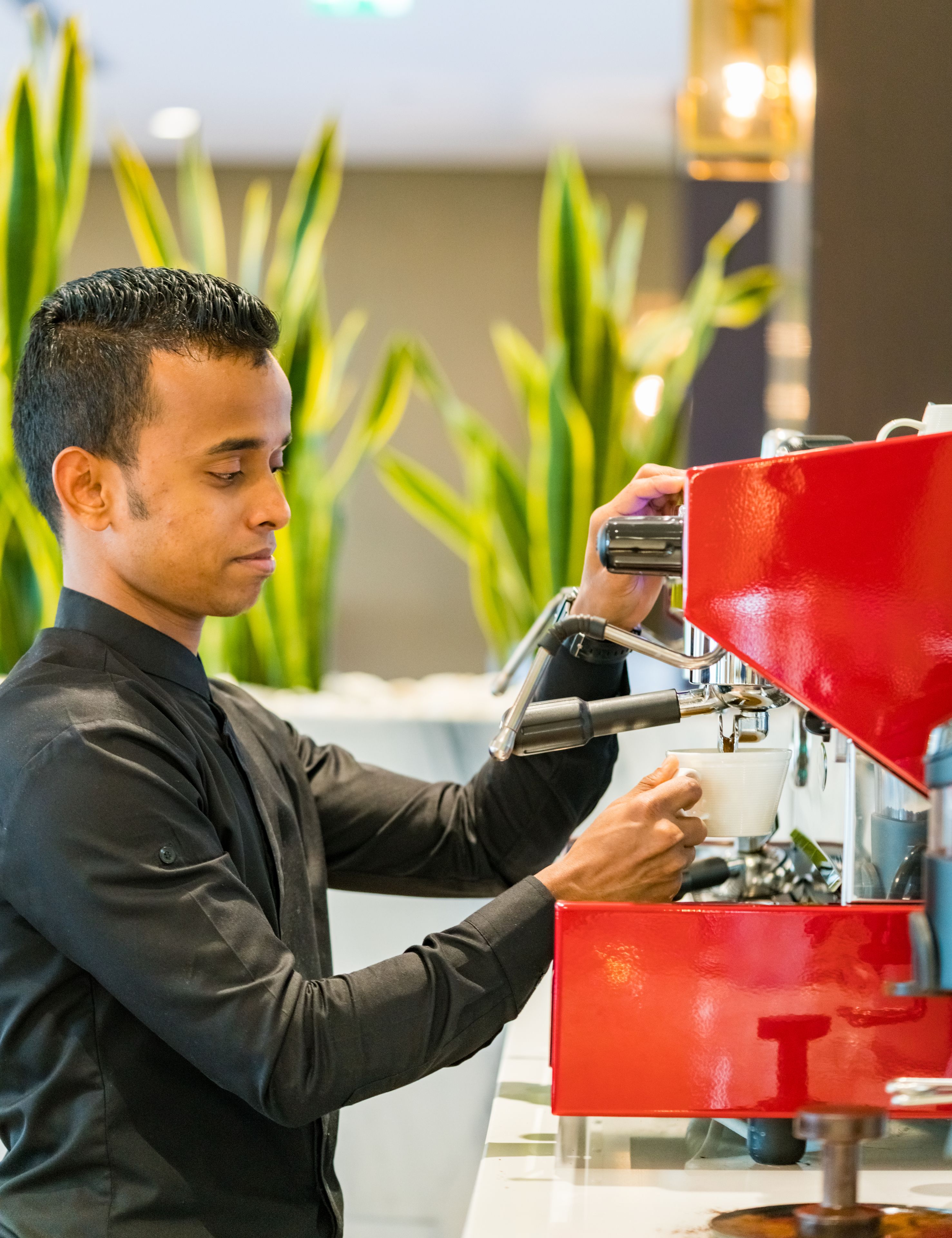 Une personne utilise une machine à expresso rouge et tient une tasse blanche à la main, dans un décor moderne avec des plantes vertes en arrière-plan.