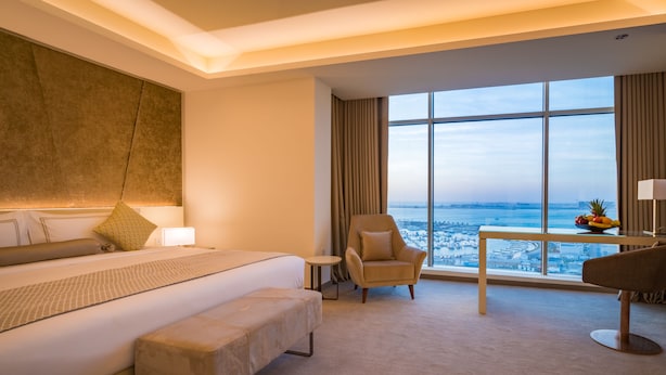 A hotel room with a large bed, a chair, a small table with fruit, and a view of the sea.