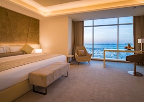 Quarto de hotel com uma cama grande, uma cadeira, uma mesinha com frutas e vista para o mar.