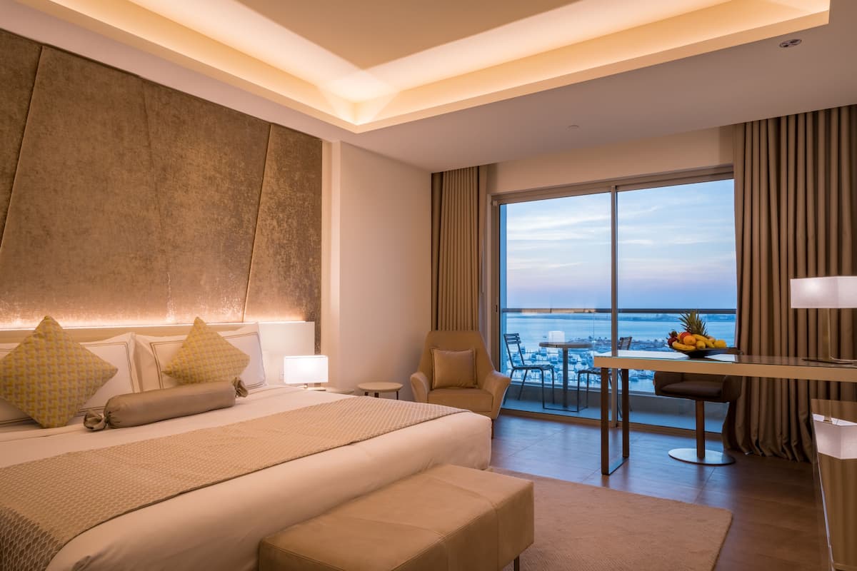 Quarto de hotel com uma cama grande, uma escrivaninha com uma cesta de frutas e vista para o oceano.