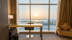 A modern hotel room with a large window, a desk, a chair, and a view of a cityscape and water.