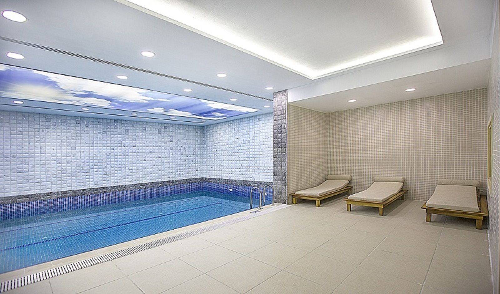 Una piscina interior moderna con pared y techo azulejados, tres sillones de descanso y una lámpara empotrada en el techo.