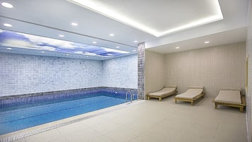 Una piscina interior moderna con pared y techo azulejados, tres sillones de descanso y una lámpara empotrada en el techo.