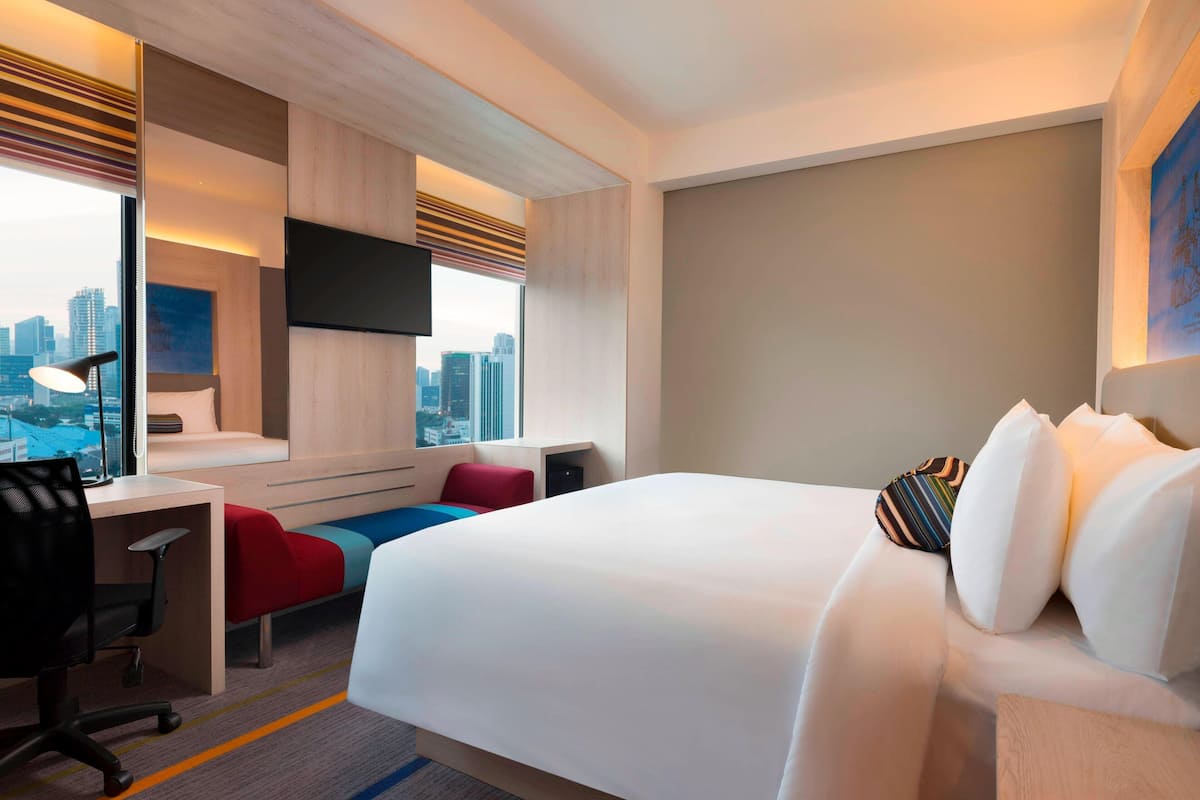 A modern hotel room with a large bed, a desk, a chair, and a view of the cityscape.