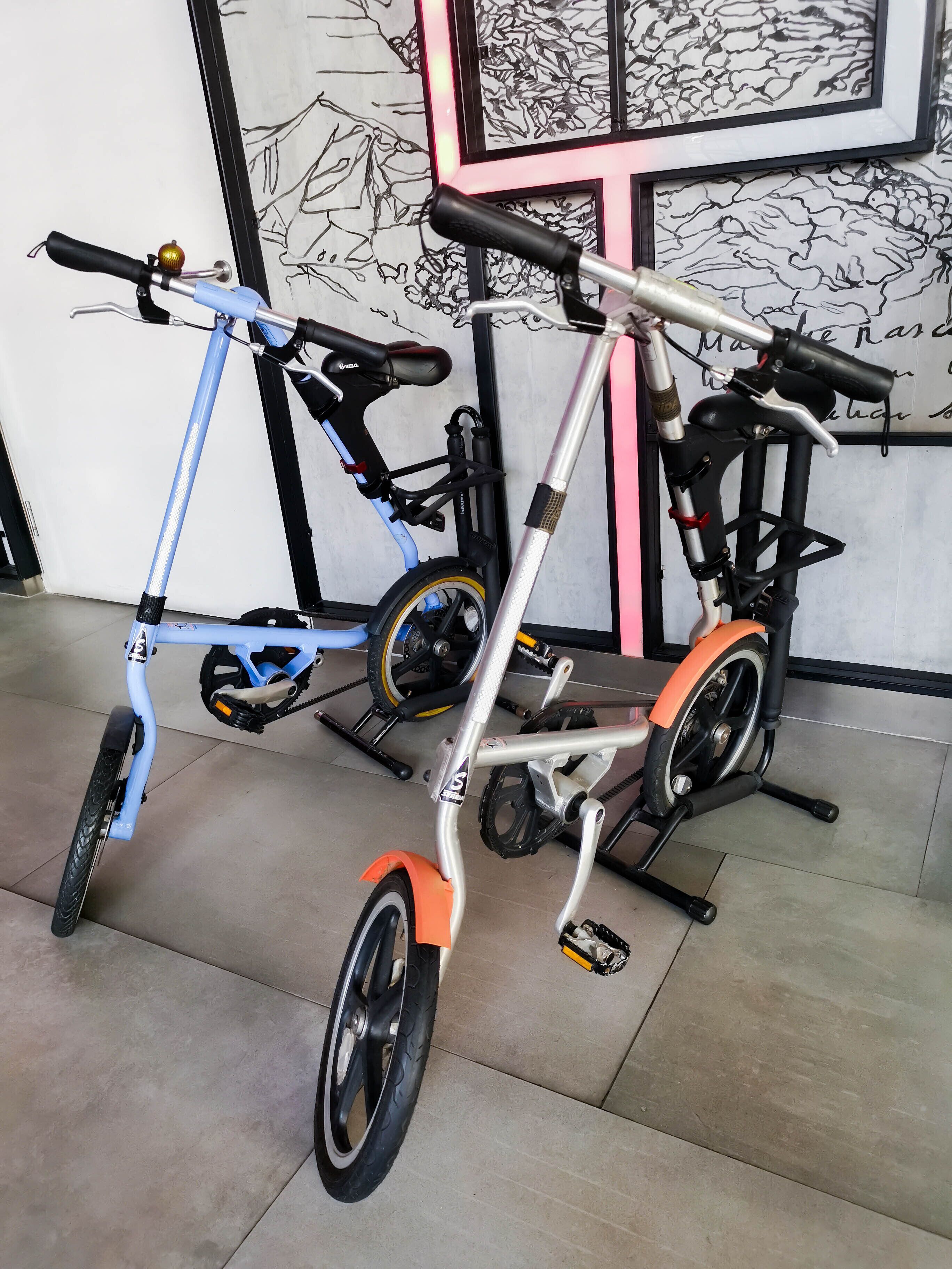En motionscykel med blå ramme og orange pedaler, placeret indendørs på et flisegulv foran en væg med et sort-hvidt abstrakt design.