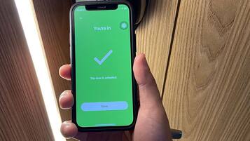 Una mano sosteniendo un teléfono inteligente que muestra una pantalla verde con una marca de verificación y el texto 'You're In' y 'The door is unlocked'.