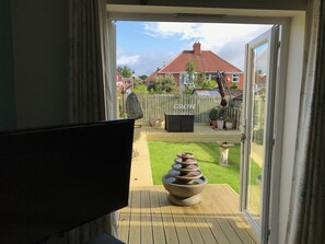 A room with a large window, a television, and a view of a backyard with a wooden deck, a bird bath, and a fence.