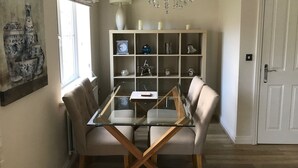 A dining area with a glass-top table, two cushioned chairs, a chandelier, a white cabinet with decorative items, and a door. - York Holiday Home (York)