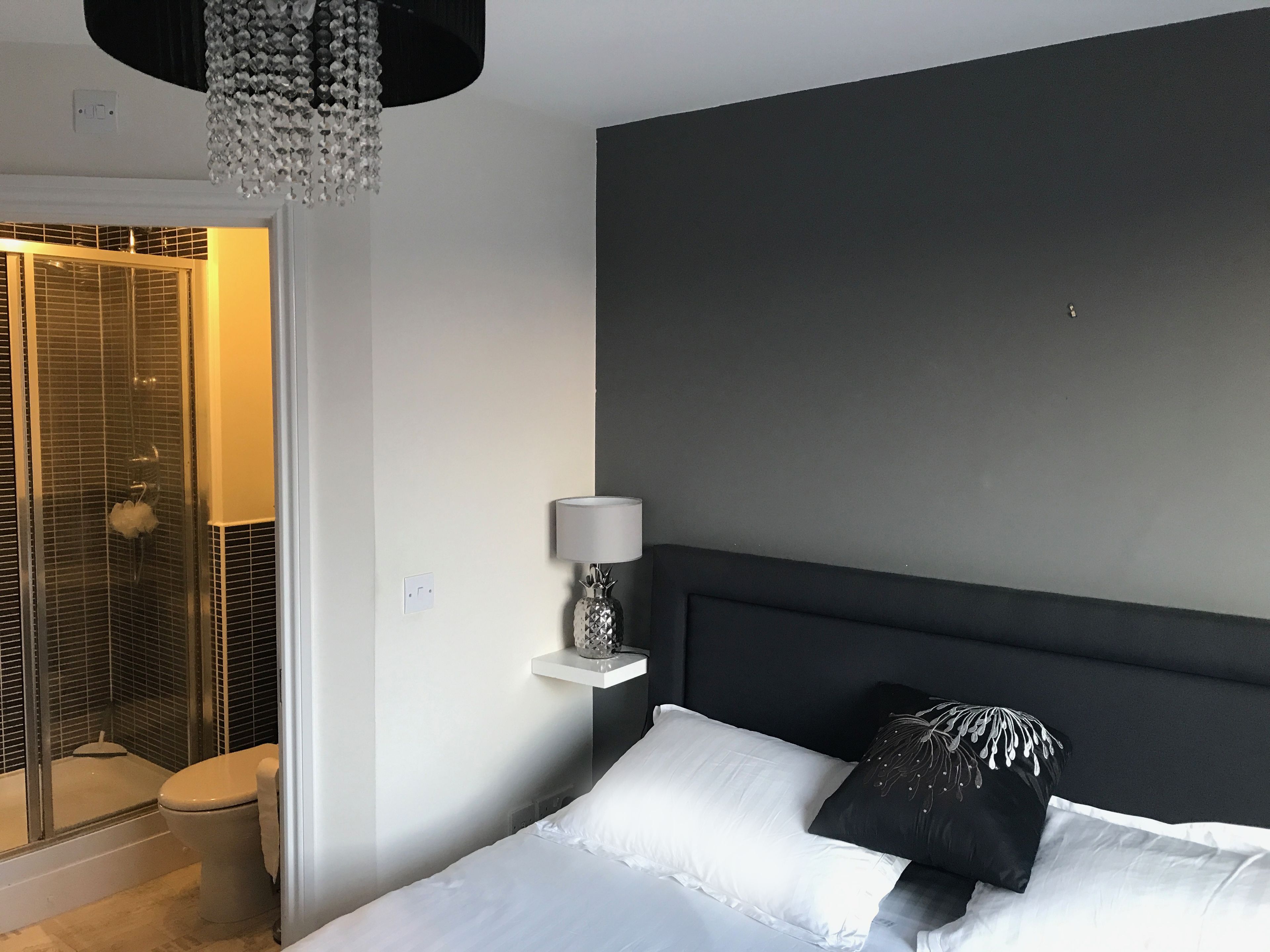 A modern bedroom with a dark headboard, white bedding, a bedside lamp, and a shower area with a glass door.