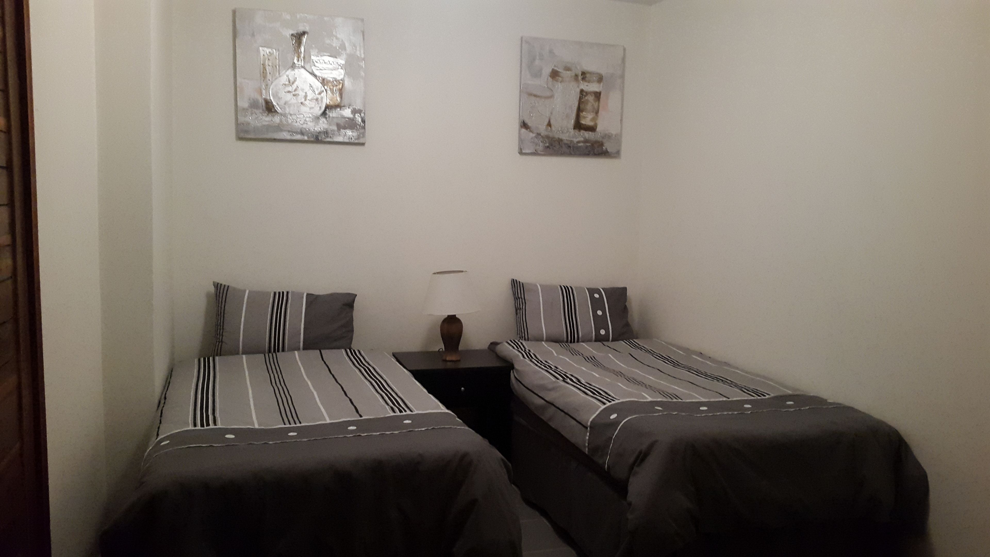 A small, simple room with two single beds, a nightstand with a lamp, and two wall-mounted paintings.