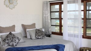 Un dormitorio con una cama grande, varios almohadones, una manta azul decorativa, un adorno en la pared y una ventana con cortinas.