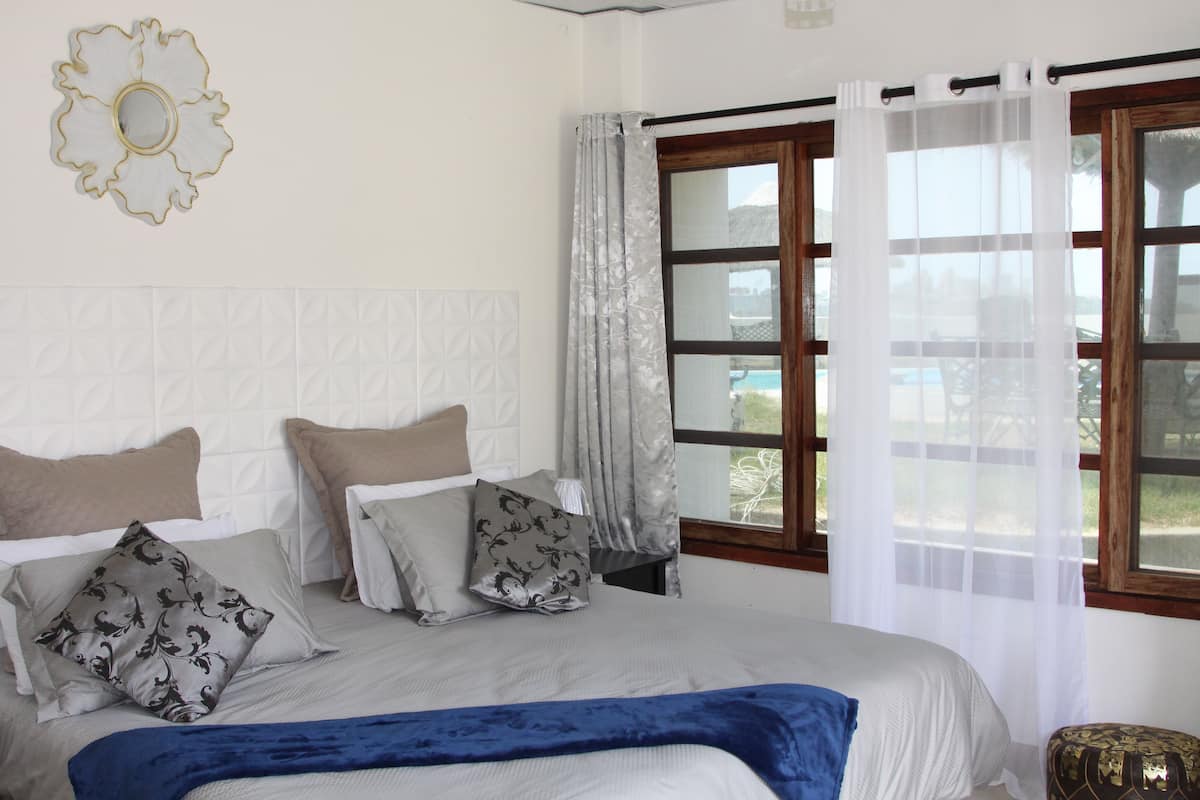 A bedroom with a large bed, multiple pillows, a blue throw blanket, a decorative wall piece, and a window with curtains.