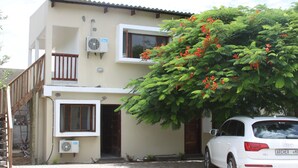 Un edificio de dos pisos con un balcón, un coche estacionado adelante y un árbol con flores rojas.