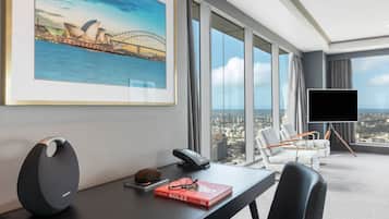 A modern hotel room with a large window offering a city view, a desk with a telephone, a framed picture of a bridge, and a flat-screen TV on a stand.