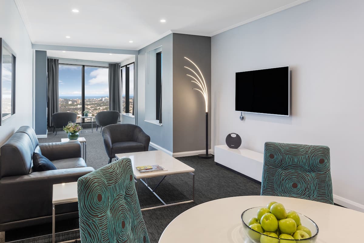 A modern hotel room with a sofa, chairs, a coffee table, a TV, and a city view.