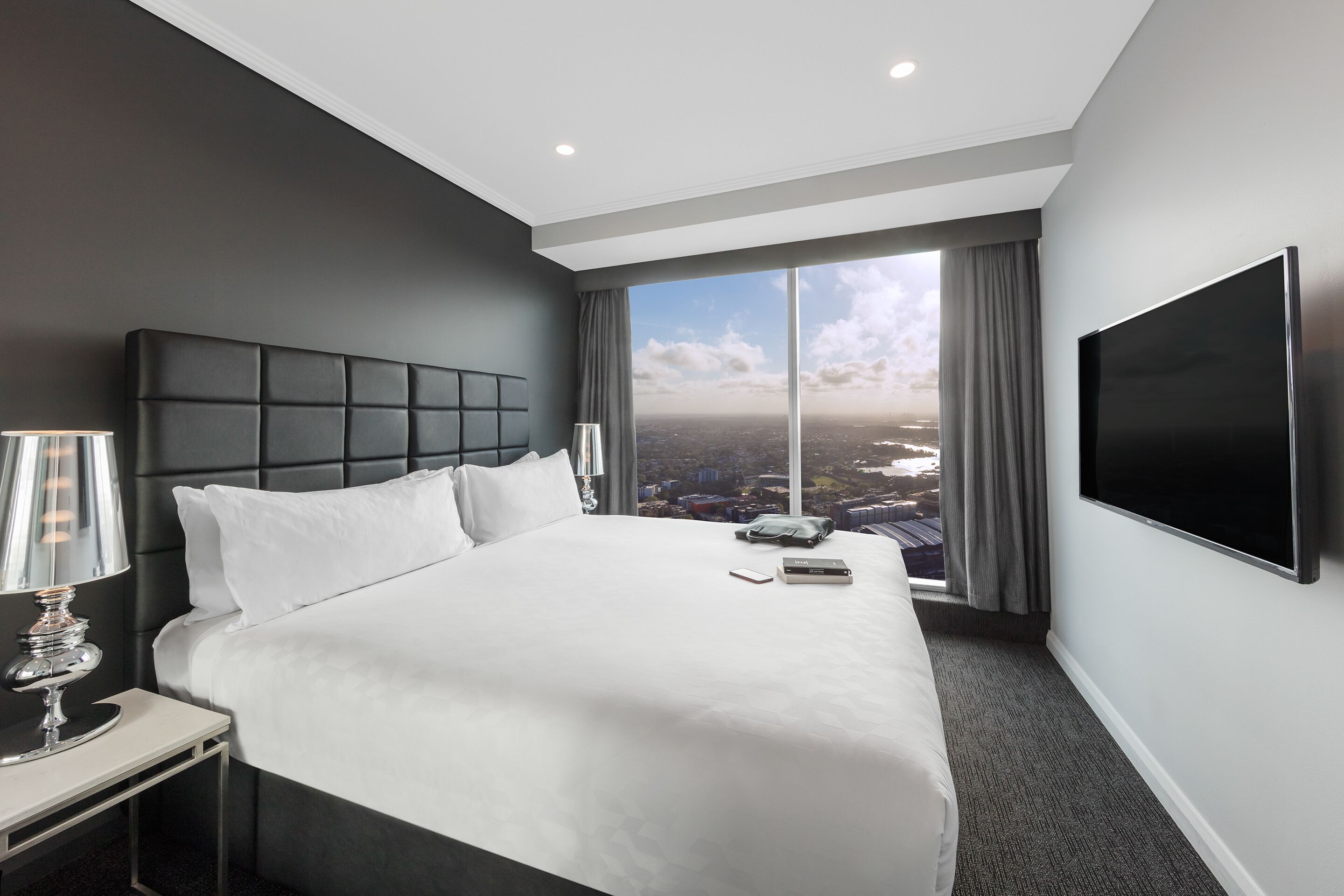Habitación de hotel moderna con una cama grande, televisor de pantalla plana, mesita de noche con lámpara y vistas a la ciudad a través de grandes ventanales.