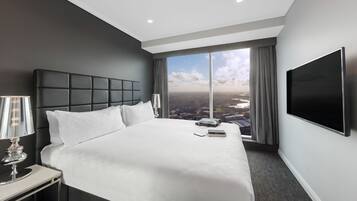 Habitación de hotel moderna con una cama grande, televisor de pantalla plana, mesita de noche con lámpara y vistas a la ciudad a través de grandes ventanales.