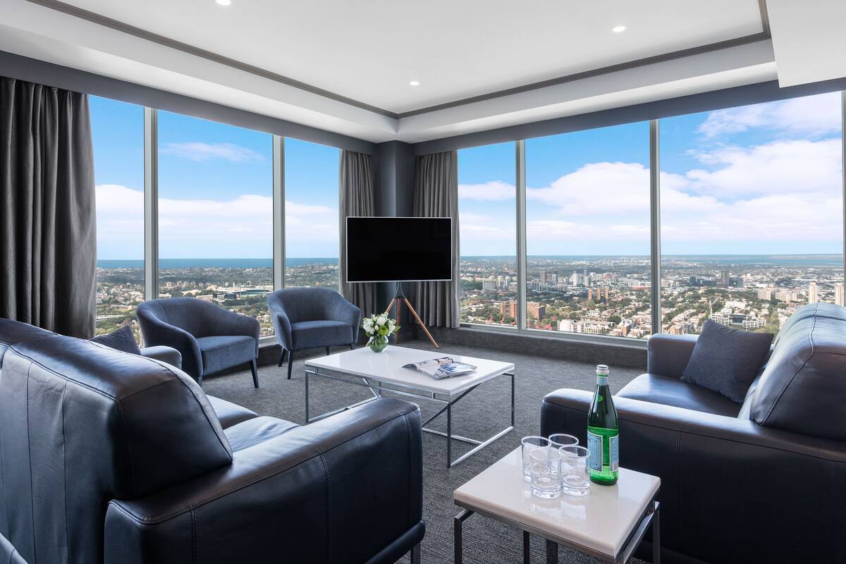 A modern hotel room with a large window, leather sofas, a coffee table, and a flat-screen TV.