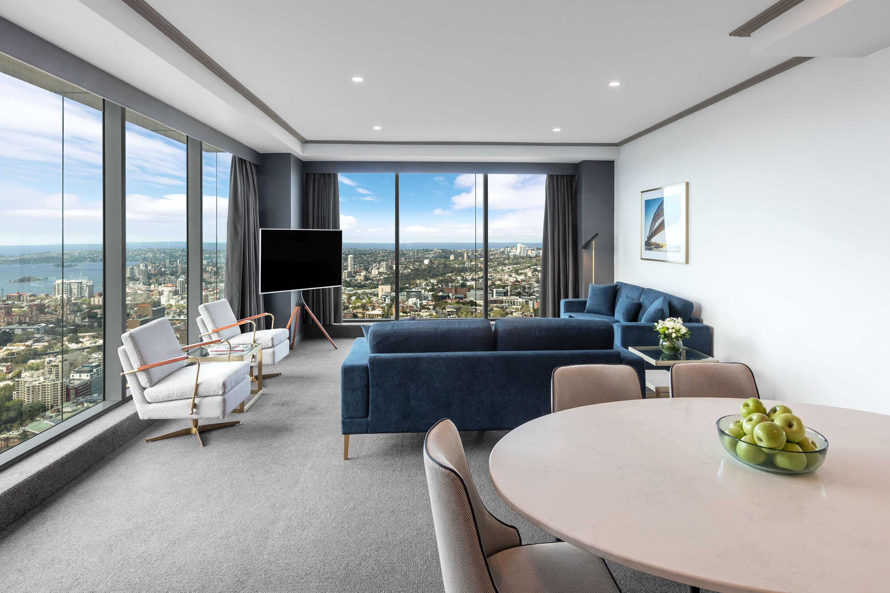 A modern living room with a large window, a blue sofa, a round table with a bowl of apples, and a flat-screen TV.