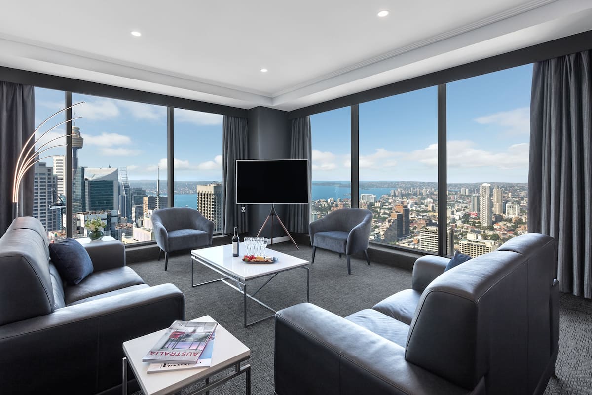 A modern hotel room with a large city view, a flat-screen TV, leather sofas, and a coffee table.