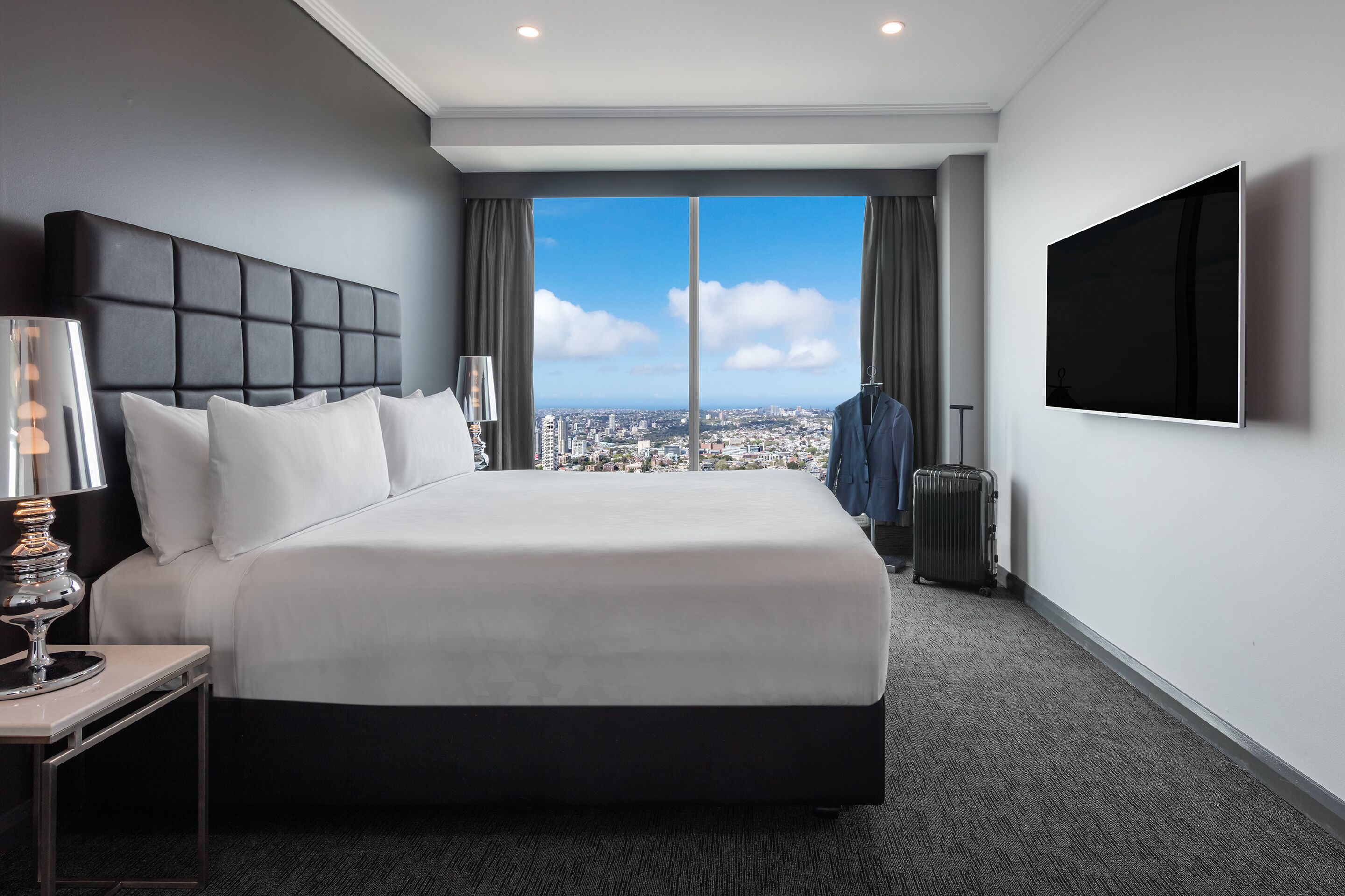 Una habitación de hotel moderna con una cama grande, un televisor de pantalla plana, vistas a la ciudad y un carrito de equipaje.