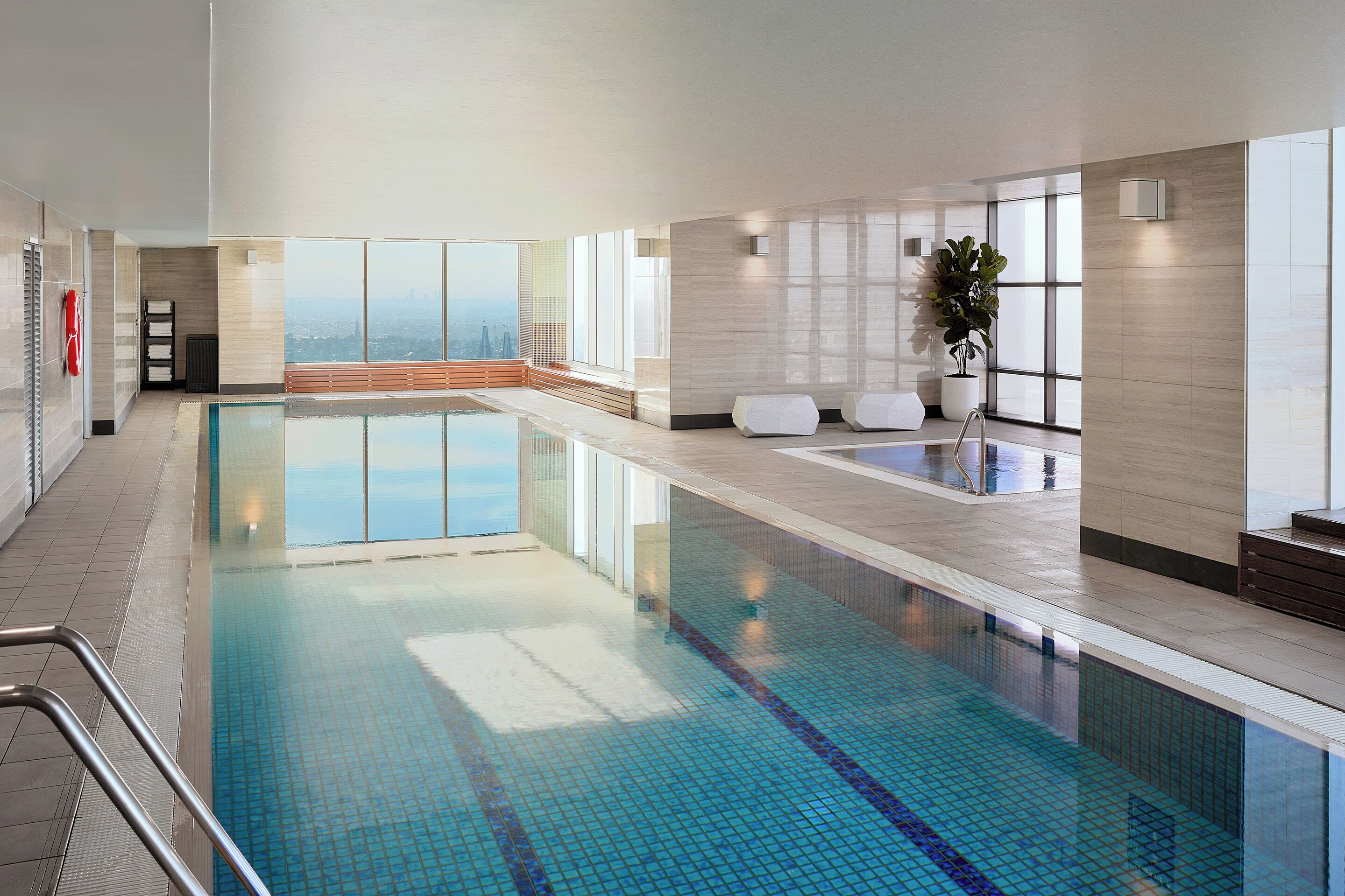 A modern indoor swimming pool with clear glass walls, a view of the cityscape, and a seating area with a potted plant.