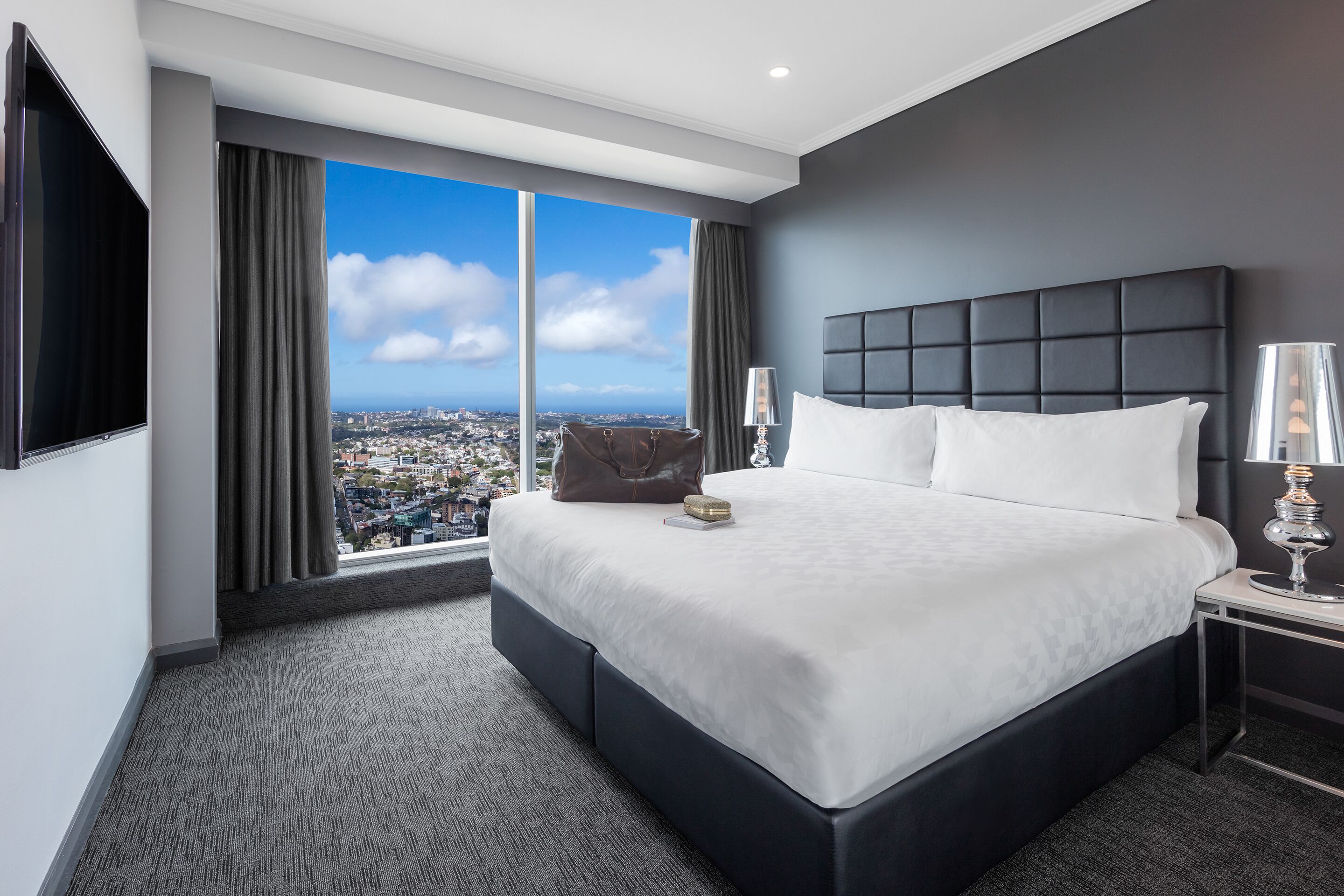 A modern hotel room with a large bed, a nightstand, a TV, and a view of a cityscape.
