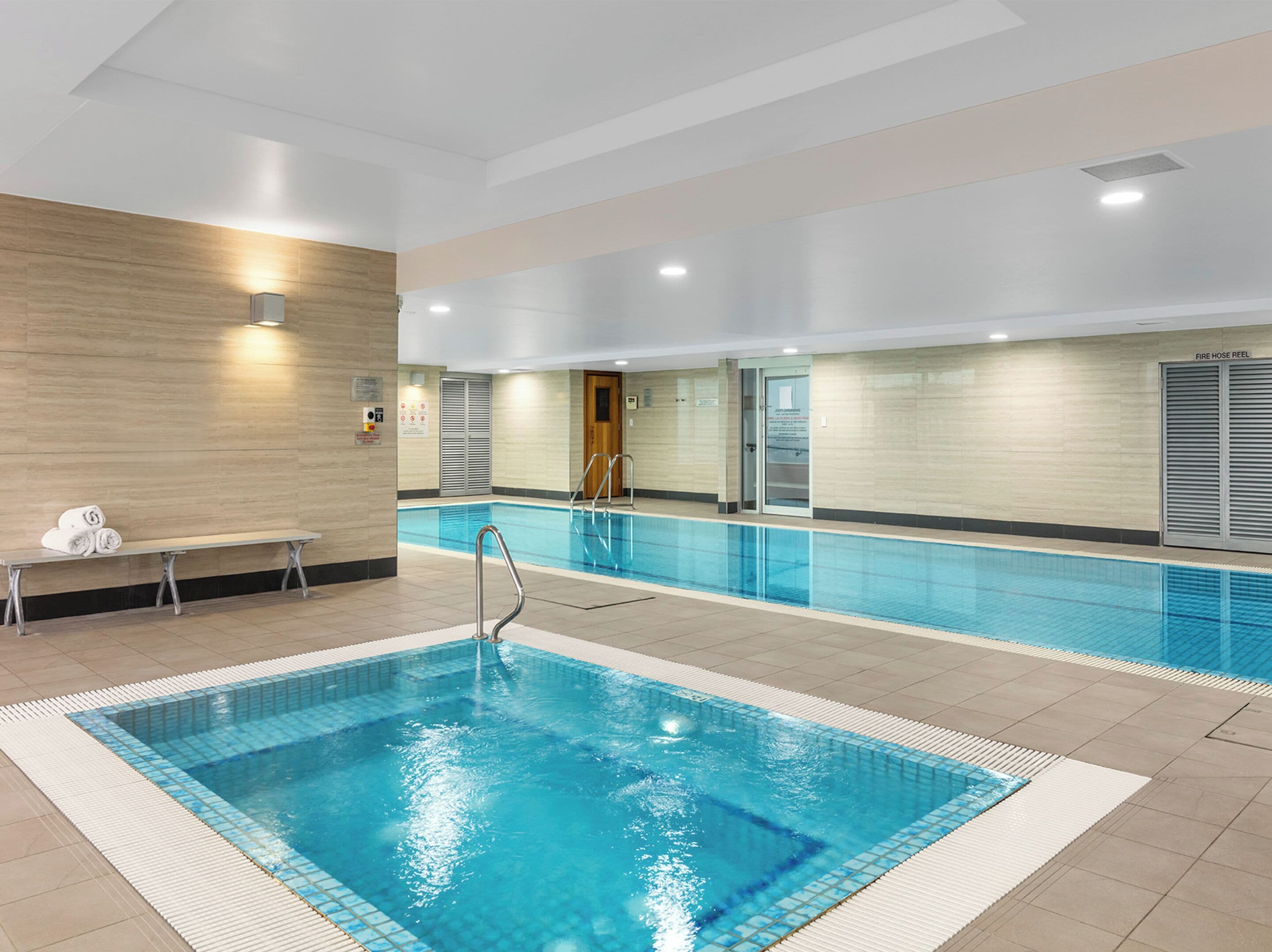 A swimming pool with a tiled deck, a bench with towels, and a fire hose reel on the wall.