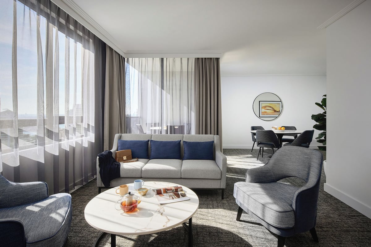 A modern hotel room with a sofa, armchairs, a round coffee table, and a dining area with a table and chairs.