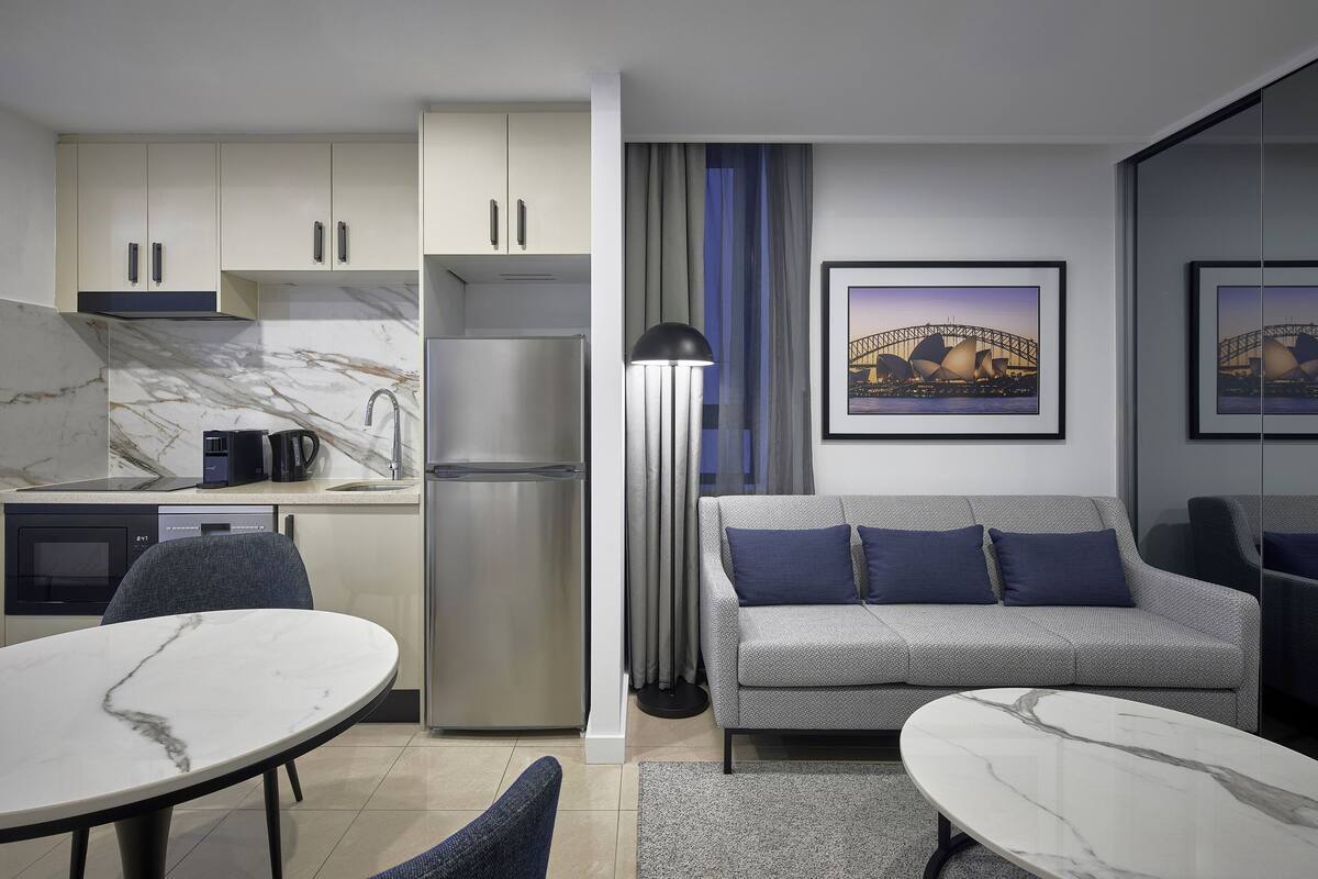 A modern kitchen with marble countertops, a built-in oven, and a refrigerator. There is a dining area with a round table and chairs, and a sofa with blue cushions.