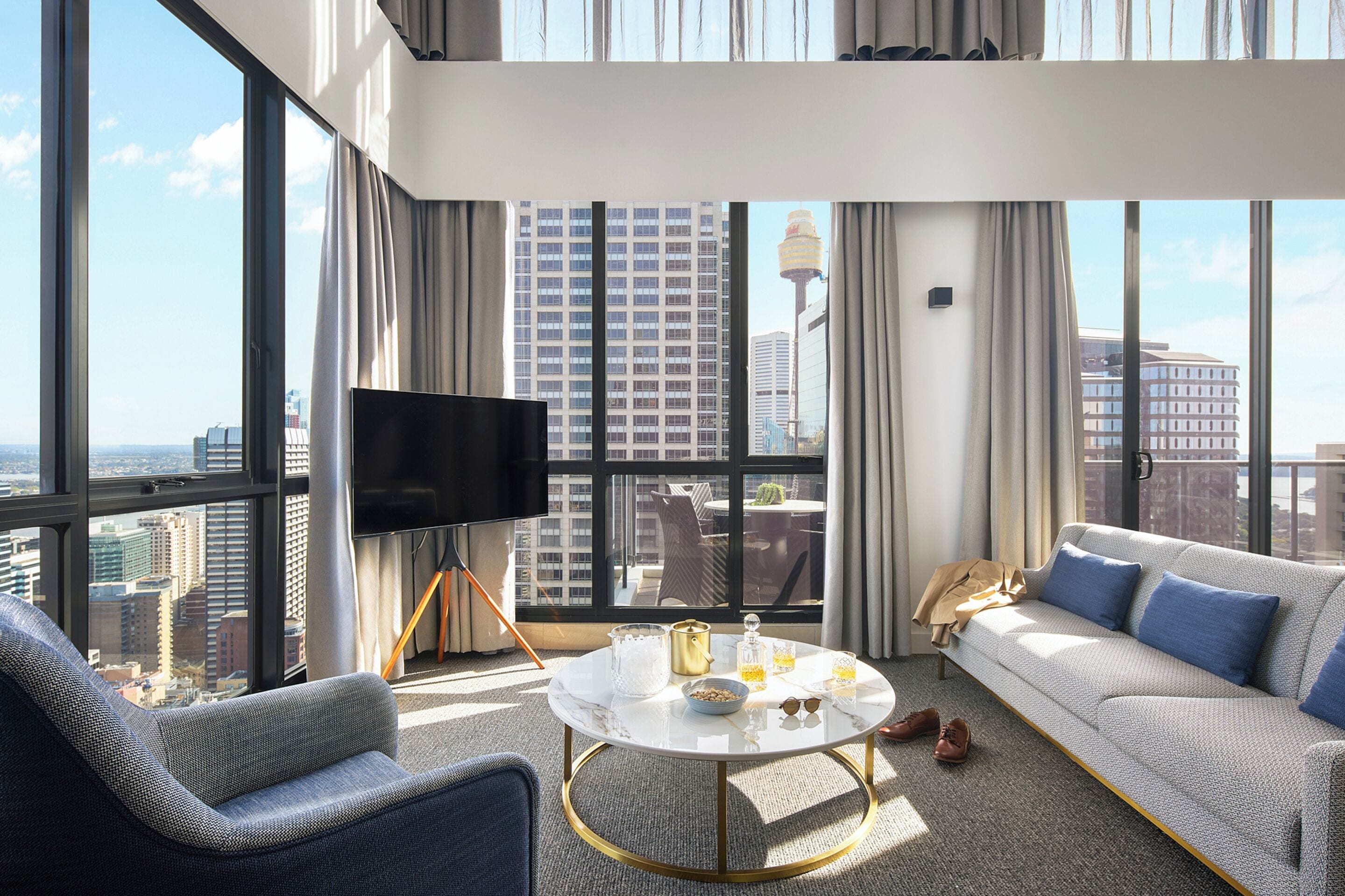 Habitación de hotel moderna con televisor grande, sofá, mesa de centro y vistas a la ciudad.