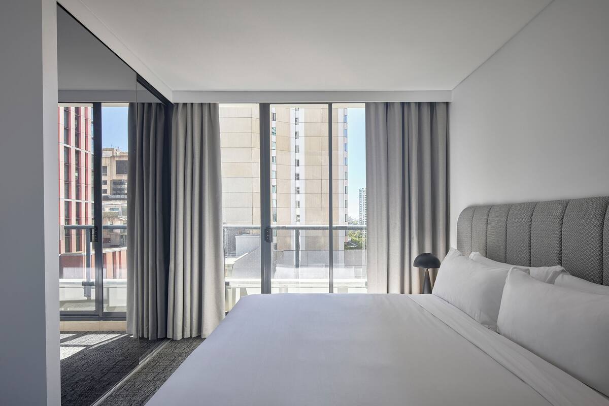 A modern hotel room with a large bed, a view of the city through floor-to-ceiling windows, and a balcony.